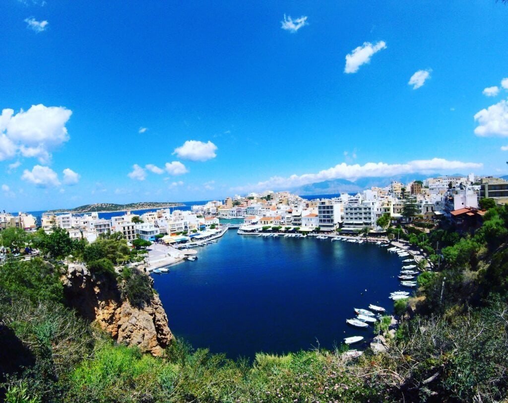 DCIM100GOPRO Coastal harbor town with white buildings along a marina, blue water and a clear blue sky with scattered clouds, taken from a rocky cliff.