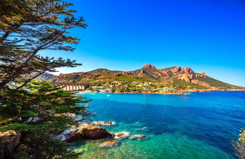 Coastal town along a bright blue sea with an arched stone bridge, rocky hills, and pastel houses under a clear sky.