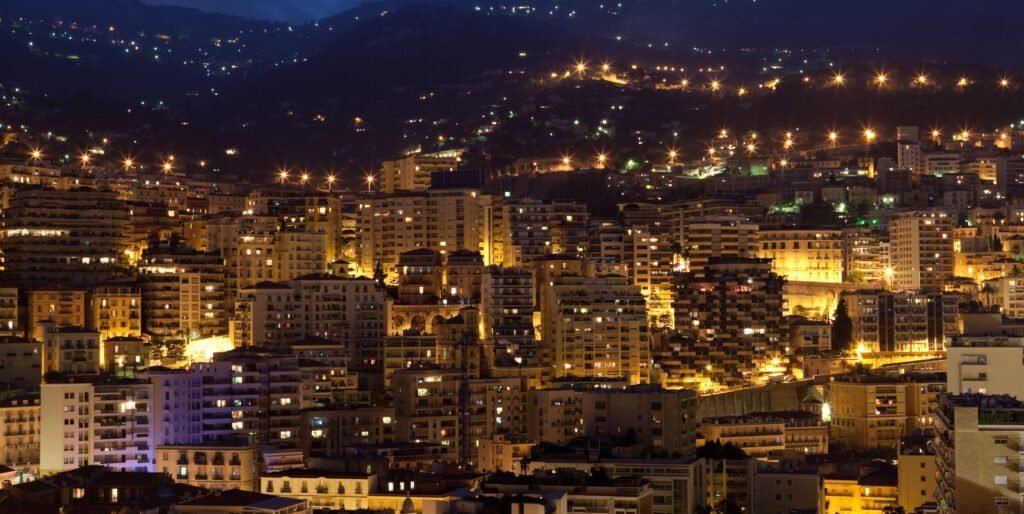 monaco-by-night-PVE7DLM Night cityscape at dusk with densely packed buildings and warm streetlights along a hillside town or cityscape.