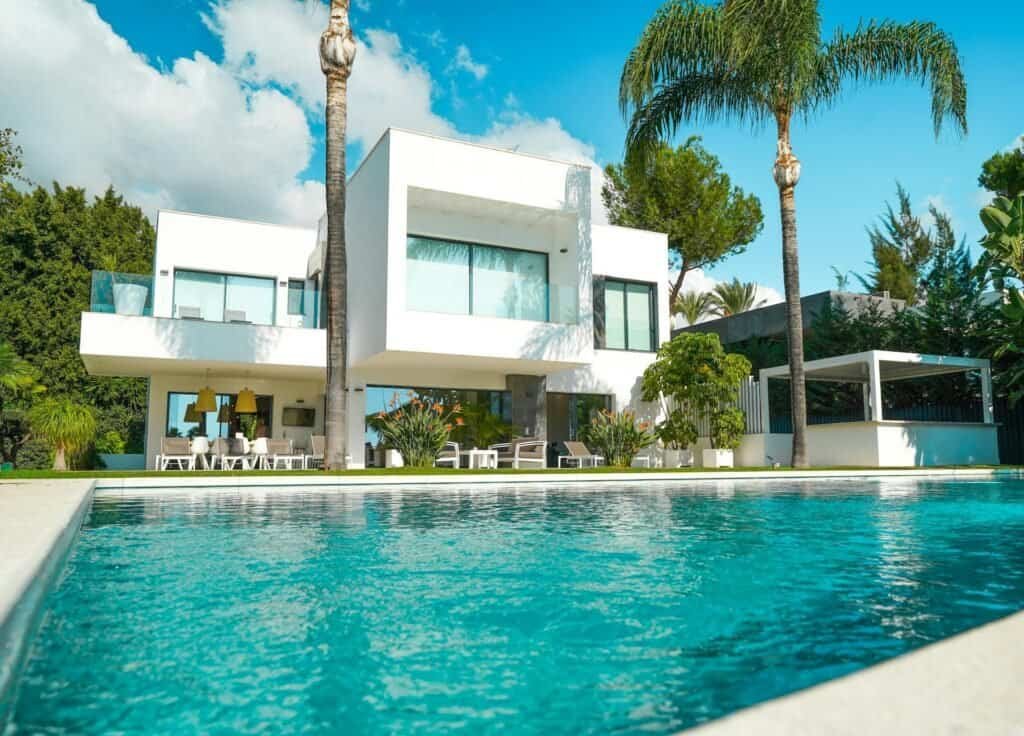 Modern white villa with large glass windows, outdoor dining area, and turquoise pool amid palm trees under a blue sky.