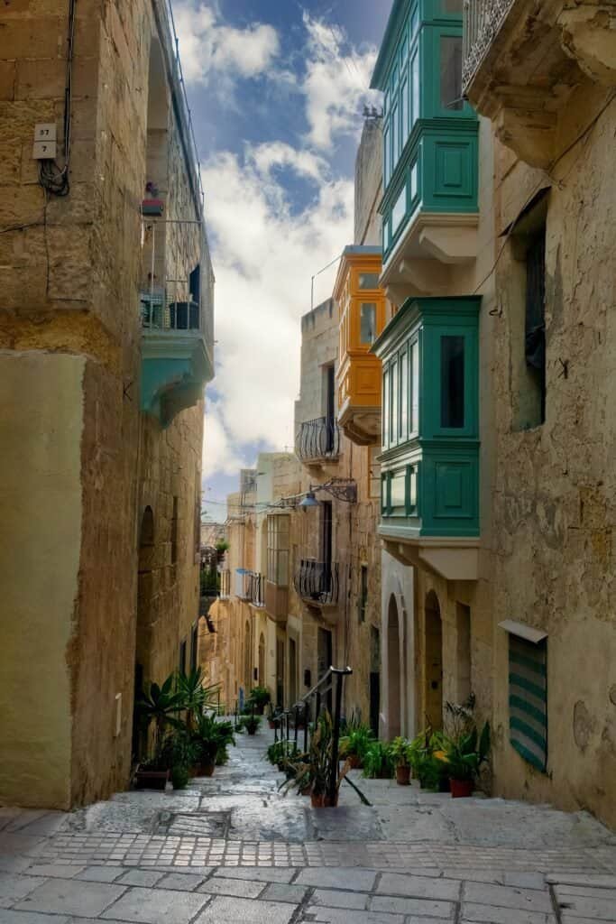 Narrow stone alleyway in a Mediterranean town, with green and yellow bay windows protruding from tall buildings and potted plants along the steps.