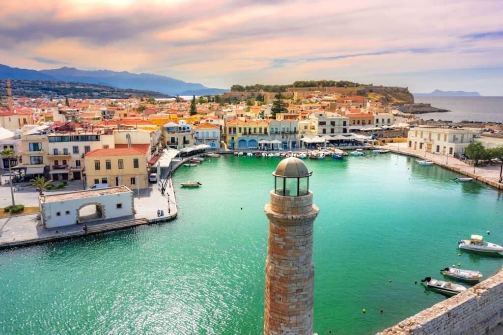 Colorful Mediterranean harbor town with pastel buildings, boats at the dock, and a tall stone lighthouse in the foreground over turquoise water