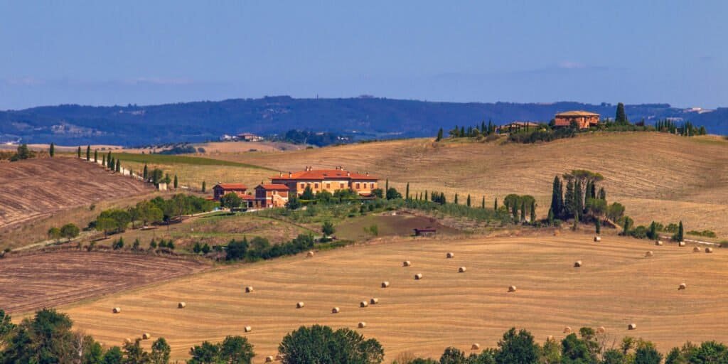 Rolling Tuscan countryside with golden fields, hay bales, and a red-roof farm surrounded by cypress trees under a blue sky